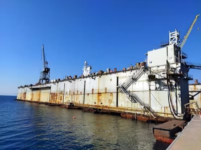 Large floating dry dock moored at a shipyard with visible rust stains, industrial equipment, and calm blue sea under a clear sky.